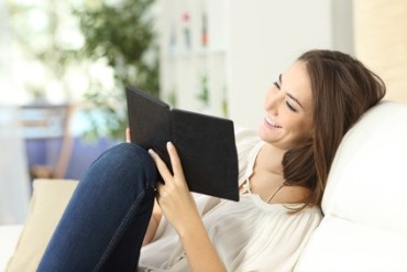 Junge Frau mit E-Book-reader sitzt auf einem wei&szlig;en Sessel in einem Zimmer