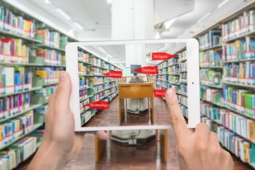 H&auml;nde, die ein Tablet halten, um einen Raum mit B&uuml;cherregalen zu fotografieren